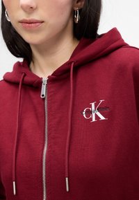 Burgundska zip-up dukserica od mekanog materijala, s kapuljačom na zatvaranje s uzicom i izvezenim 'Calvin Klein' logotipom u crnoj i bijeloj boji.