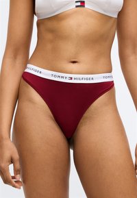 Tumši sarkani stringi ar baltu jostu ar uzrakstu "TOMMY HILFIGER." Mīksta auduma ar gludu tekstūru un minimālas sedzības dizains.