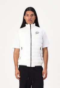 Balta pūkinė liemenė su aukštu apykakle ir užtrauktuku, su juodais užtrauktukais kišenėse ir juodu logotipu ant krūtinės. Tekstūruotas siuvinėjimo dizainas.