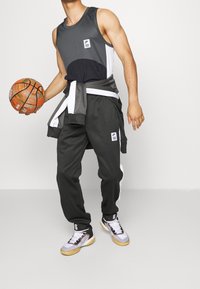 Tenue de sport composée d'un débardeur gris, d'un pantalon de survêtement noir et gris avec des accents blancs, et de baskets de basketball noires, tenant un ballon de basketball orange.