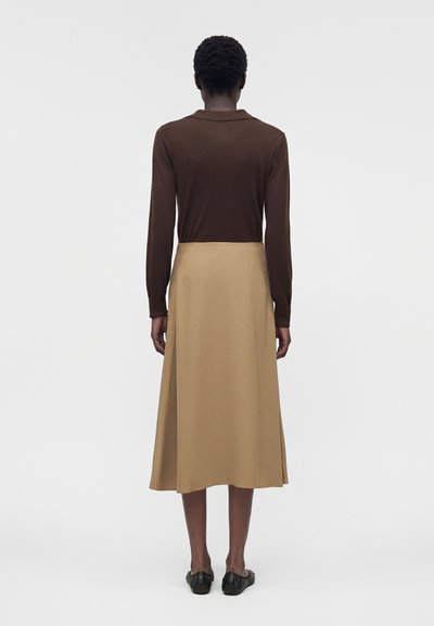 Άτομο που φορά καφέ μπλούζα με μακριά μανίκια, μπεζ midi φούστα και μαύρα παπούτσια, στέκεται γυρισμένο με την πλάτη σε μονόχρωμο λευκό φόντο.
