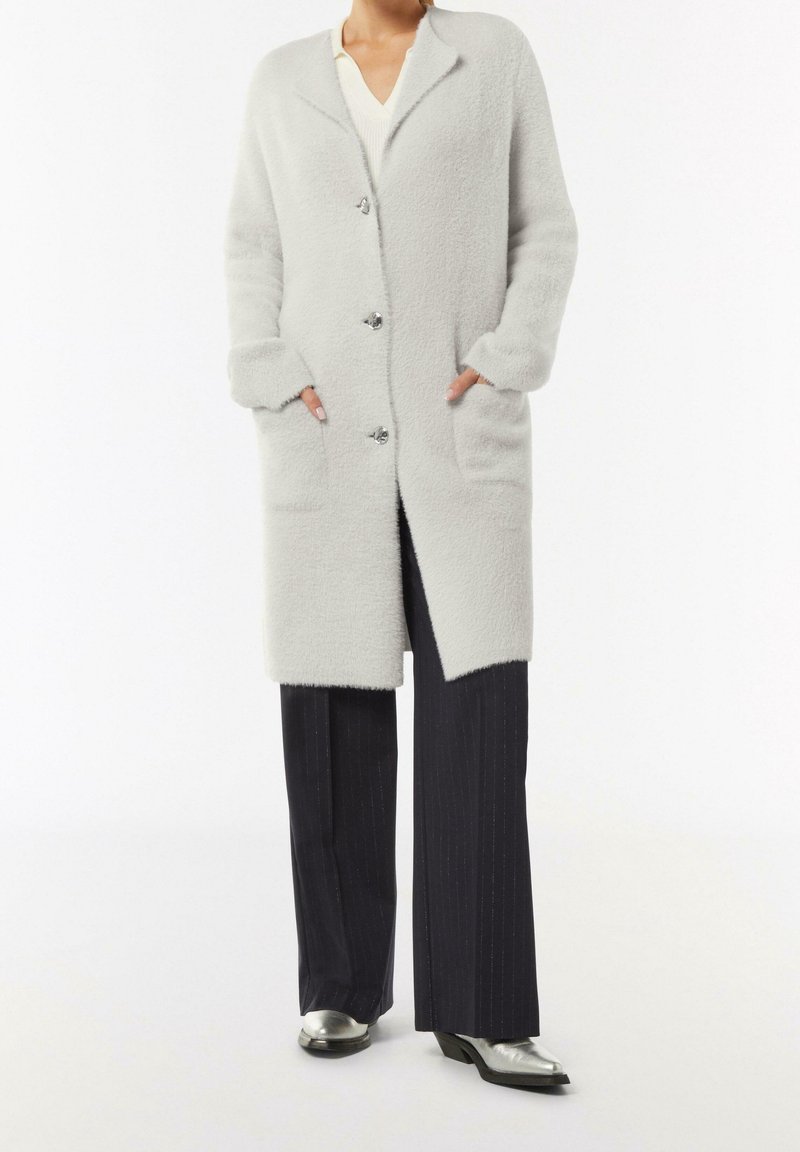 Femme portant un long manteau gris clair texturé avec des boutons, un pantalon noir à jambes larges et des chaussures argentées à bout pointu, debout les mains dans les poches.