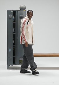 Witte sportjas met rode strepen, grijze joggingbroek en zwarte schoenen. Kluisje en houten bank op de achtergrond, basketbal bovenop het kluisje.