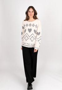 Strickpullover in Creme mit grauen Herz- und Diamantmustern, kombiniert mit schwarzen weit geschnittenen Hosen. Besitzt einen lässigen Schnitt und einen Rundhalsausschnitt.