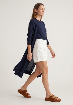 Kvinde går iført en navy lang cardigan, navy top, hvid nederdel og brune platformsandaler mod en lys ensfarvet baggrund.