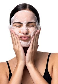 Tuchmaske auf einem Gesicht, aus dünnem Stoff. Die Maske ist weiß mit Ausschnitten für Augen und Mund. Die Haut wirkt glatt und die Hände sind sanft auf die Wangen gedrückt.