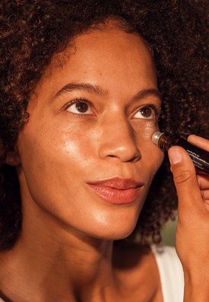 Rollerball serum applicator with brown glass bottle; model has curly hair, applying product under her eye; natural skin texture and glow.