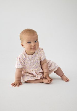 Baby with light hair and blue eyes sitting on white floor wearing a short-sleeve lavender dress with small floral print.