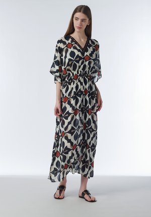 Vrouw draagt een zwarte, witte en rode losse maxi-jurk met ellebooglange mouwen en zwarte sandalen, staand tegen een effen achtergrond.