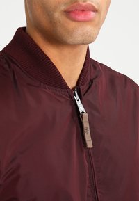 Jachetă bomber burgundy, cu guler ribbit, închidere cu fermoar și textură netedă, lucioasă. Detalii notabile includ un fermoar cu logo.