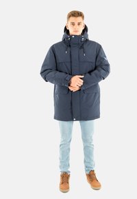 Parka impermeabile blu navy con colletto alto, cappuccio regolabile, grandi tasche frontali e chiusura frontale con bottoni. Jeans azzurri e stivali marroni.