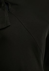 Tissu noir avec une couture diagonale et un détail de nœud noué près de l'encolure, montrant la texture et les coutures de près.