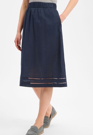 Vrouw draagt een marineblauwe knielange rok met elastische taille en decoratieve zoomdetails, gecombineerd met grijze instapschoenen.