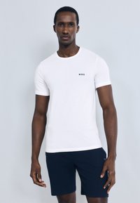 Bílé tričko s kulatým výstřihem z bavlny, na hrudi s malým tmavým logem. Kombinováno s tmavě modrými šortkami. Hladká textura.