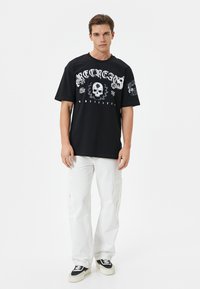 Schwarzes Baumwoll-T-Shirt mit grafischen Drucken, die ein Skull, florale Elemente und Text zeigen. Getragen mit weißen Cargo-Hosen und schwarzen Sneakers.