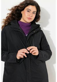 Zwarte gewatteerde jas met hoge kraag en verstelbare trekkoorden, voorzien van een ritssluiting en een relaxed fit. Draag het over een paarse trui.