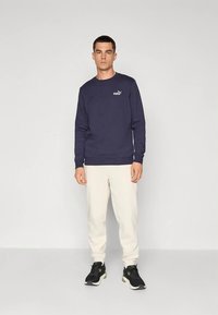 Sudadera azul marino de algodón con cuello redondo, mangas largas y logo blanco de Puma. Combinada con pantalones crema con cordón y zapatos deportivos negros.