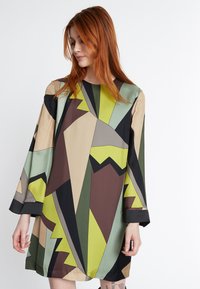 Robe à motifs géométriques présentant un mélange de verts, marrons et beiges. Le design inclut des formes angulaires et de longues manches avec un ourlet noir.