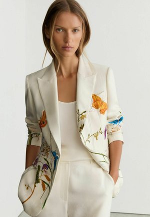 Femme portant un costume blanc avec des imprimés floraux et papillons colorés, mains dans les poches, et un top blanc sans manches en dessous.