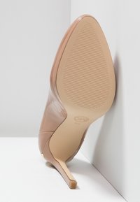 Chaussure à talon aigu en cuir verni nude avec un bout pointu et un talon stiletto mince. La semelle présente des rainures horizontales pour une meilleure adhérence.