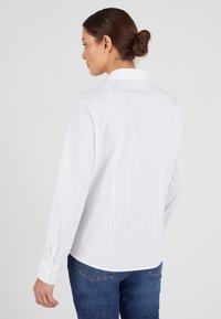Camisa blanca de botones con mangas largas y cuello puntiagudo. Tela suave, corte ajustado, detalles de costura visibles, llevada con jeans de mezclilla azul.