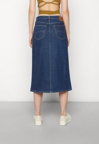 Tummansininen denim-midi-hame suoralinjaisella leikkauksella, jossa on kaksi takataskua ja etiketti vyötäröllä. Yhdistetty valkoisiin lenkkareihin.