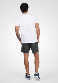 T-shirt blanc à manches courtes associé à un short gris foncé, porté avec des chaussures de sport bleues et blanches. Tatouage visible sur le bras gauche.
