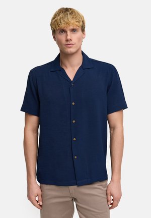 Homme blond portant une chemise bleu marine à manches courtes boutonnée et un pantalon beige, debout devant un fond clair uni.
