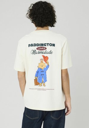 T-shirt crème avec un graphisme de Paddington Bear portant un manteau bleu, tenant un chapeau et une valise. Le texte indique "Paddington aime la marmelade."