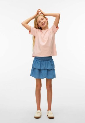 Joven niña con una camiseta rosa y una falda azul de capas está de pie con las manos levantadas por encima de su cabeza, sonriendo, sobre un fondo blanco liso.