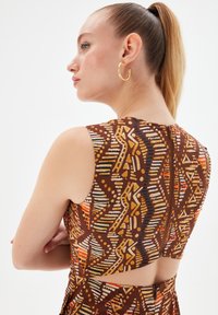 Vestido marrón con estampado, de diseño sin mangas, que presenta impresiones geométricas en naranja y amarillo. Espalda descubierta y un detalle de cremallera.