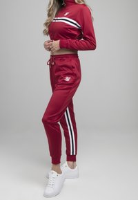 Kvinde i rød træningsdragt med hvide og sorte striber, iført hvide sneakers, stående mod en ensfarvet grå baggrund.