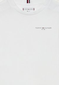 Valge puuvillane T-särk lühikeste varrukatega, mille esiküljel on väike must logo "TOMMY HILFIGER EST. 1985" ja triibuline kaelus.
