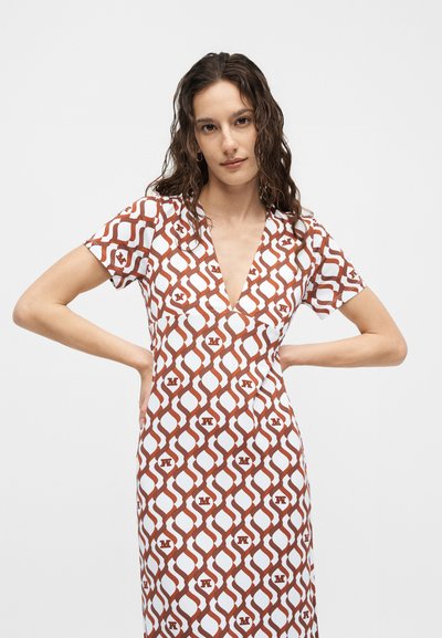 Νεαρή γυναίκα με μακριά σγουρά μαλλιά που φορά ένα καφέ και λευκό φόρεμα με μοτίβο, κοντά μανίκια και βαθύ ντεκολτέ σε σχήμα V, στέκεται με τα χέρια στη μέση.