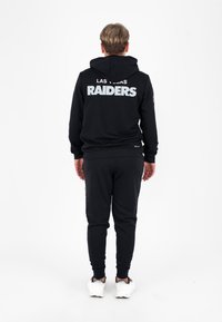 Zwarte hoodie met "LAS VEGAS RAIDERS" in vette, reflecterende letters op de achterkant. Gecombineerd met bijpassende zwarte joggingbroeken en witte sneakers.
