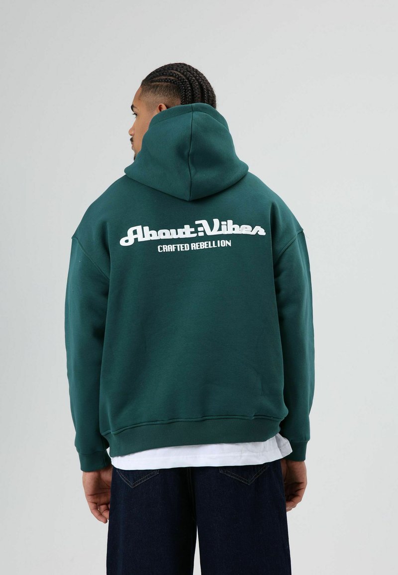 Donkergroene hoodie met een kangoeroezak, met witte tekst op de achterkant: 'About Vibes Crafted Rebellion'. Gemaakt van een katoenmix.