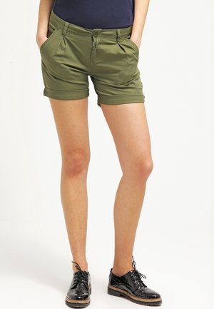 Oliwkowe szorty z podwiniętymi mankietami, z przodu kieszenie i zapięcie na zamek. W zestawieniu z czarnymi lakierowanymi butami na sznurówki.