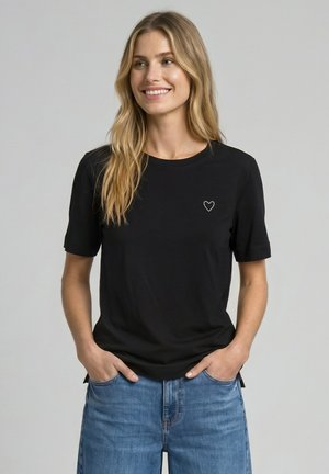 Uśmiechnięta kobieta z długimi blond włosami, ubrana w czarną koszulkę z małym białym serduszkiem i niebieskie dżinsy, ręce w kieszeniach, jasnoszare tło.