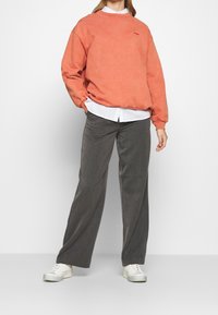 Sudadera color coral con puños y cuello acanalados, llevada sobre una camisa blanca, combinada con pantalones de pierna ancha grises y zapatillas blancas.