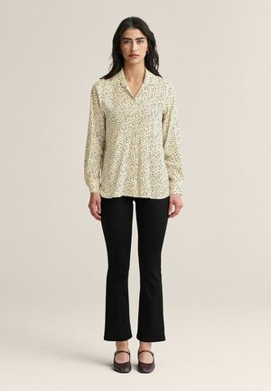 Kvinde stående iført en cremefarvet bluse med lange ærmer og sorte prikker, sorte udvidede bukser og mørke Mary Jane-sko, foran en ensfarvet baggrund.