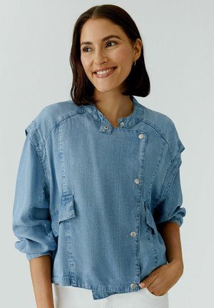Frau trägt eine hellblaue, geknöpfte Jeansjacke mit hochgekrempelten Ärmeln, lächelt und blickt seitlich vor einem einfarbigen Hintergrund.
