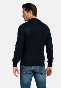 Man met kort donker haar, gekleed in een donkere kabelgebreide trui en blauwe jeans, staand met zijn rug naar de camera tegen een witte achtergrond.