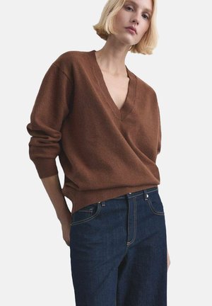 Persona que lleva un suéter marrón de cuello en V con mangas largas y jeans azul oscuro, de pie contra un fondo claro y liso.