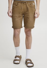 Pantaloni scurți de denim maro cu tivuri rulate, talie elastică cu șnur, buzunare frontale, purtați cu șosete albe și sandale maro.