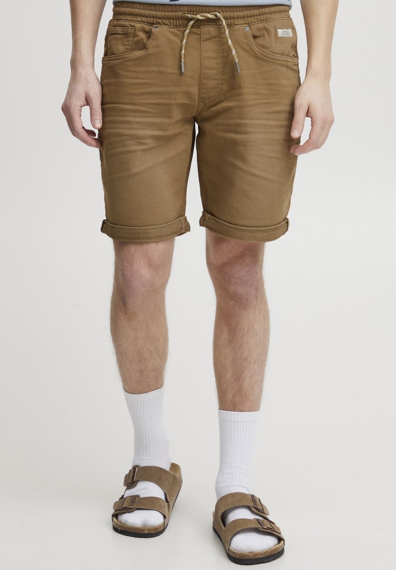 Pantaloni scurți de denim maro cu tivuri rulate, talie elastică cu șnur, buzunare frontale, purtați cu șosete albe și sandale maro.