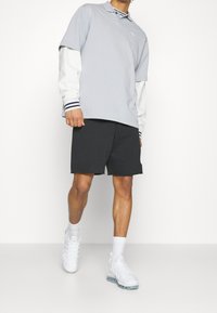 Ljusgrå polo med blå detaljer, lager på en vit långärmad tröja, i kombination med svarta shorts och vita sportskor.