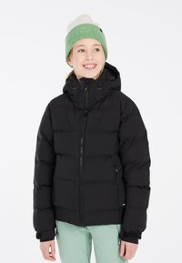 Giacca a vento nera con zip frontale, cappuccio e polsini elasticizzati. Indossata con pantaloni verde menta. Gli accessori includono un berretto verde e bianco.