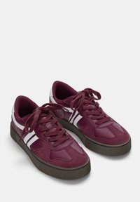 Baskets bordeaux avec des rayures blanches, présentant un mélange de matériaux lisses et texturés, bout rond et semelle en caoutchouc robuste.