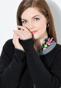 Jeune femme aux yeux bleus et aux cheveux châtains clairs portant un pull noir avec un col fleuri, tenant délicatement ses mains près de son visage.