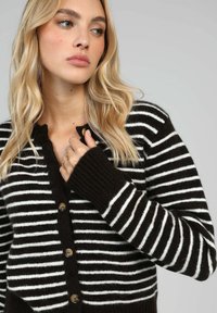 Gilet rayé en marron foncé et blanc, doté d'une fermeture à boutons, de poignets côtelés et d'une texture en maille douce. Présenté avec un col rond.